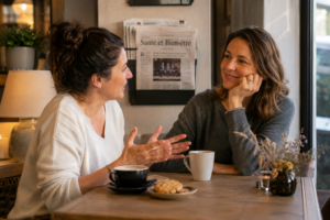 Une femme confiante, assise dans un café tranquille, discutant avec une amie, avec des tasses de thé sur la table, et un sourire rassuré.